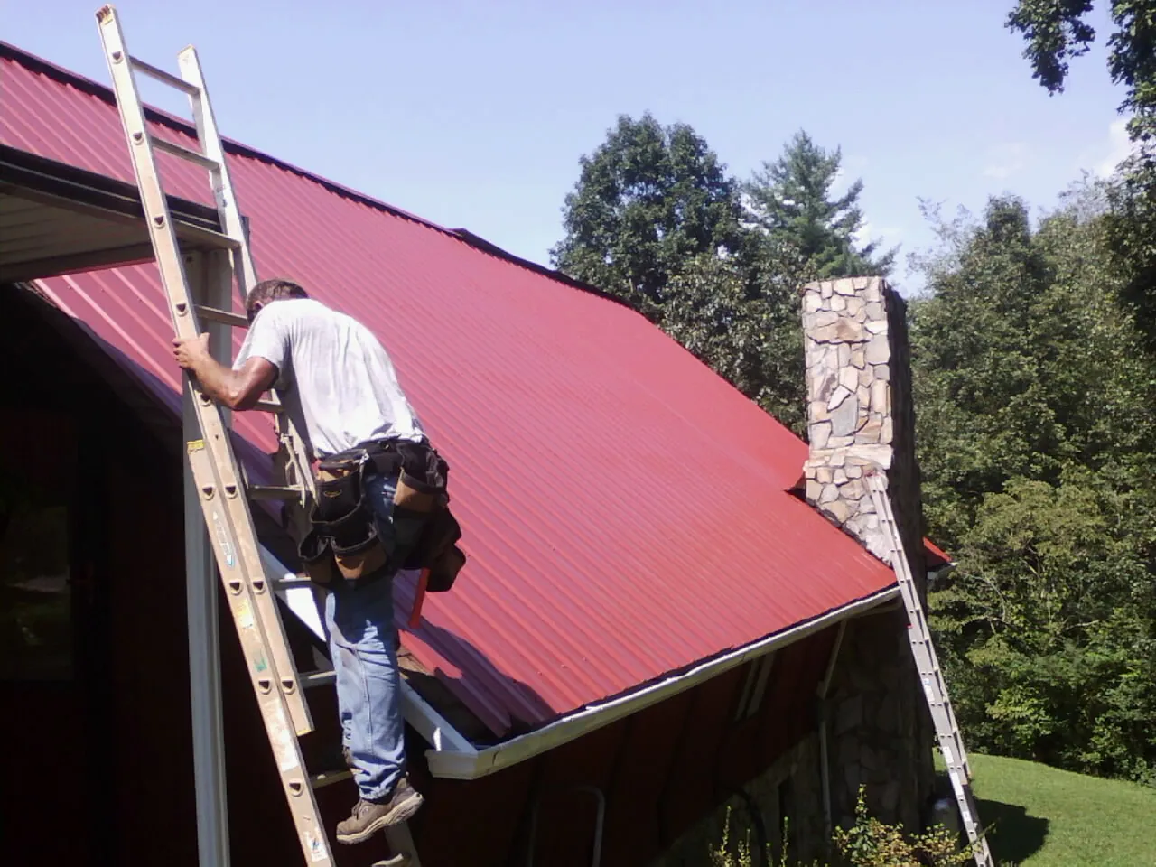 Roofer on a ladder beside a red metal roof and stone chimney — Soffit & Fascia Repair by Roofing Brothers in Westbrook